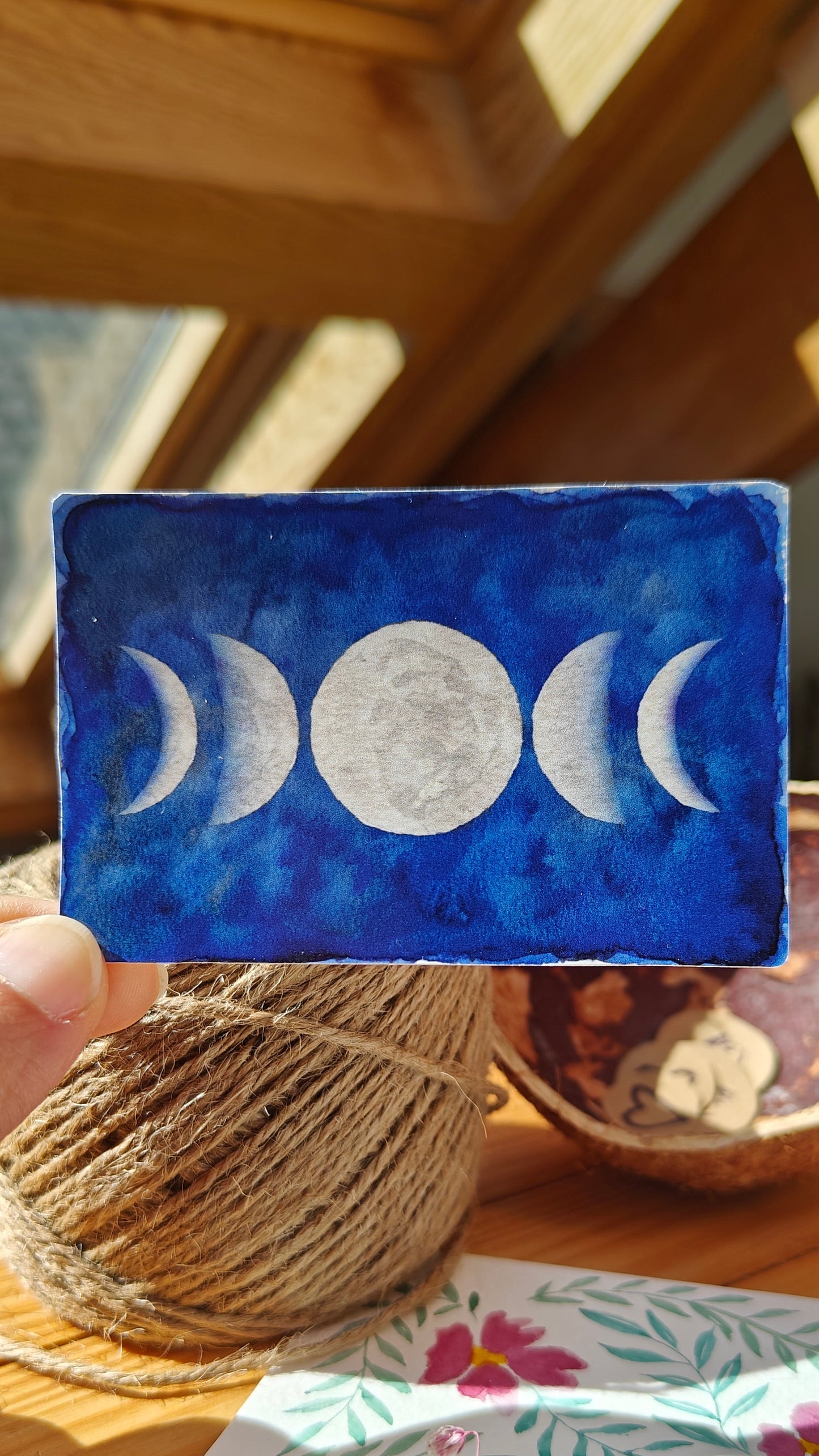 Handheld sticker with moon phases over a wooden surface with twine, watercolour painting and flowers.