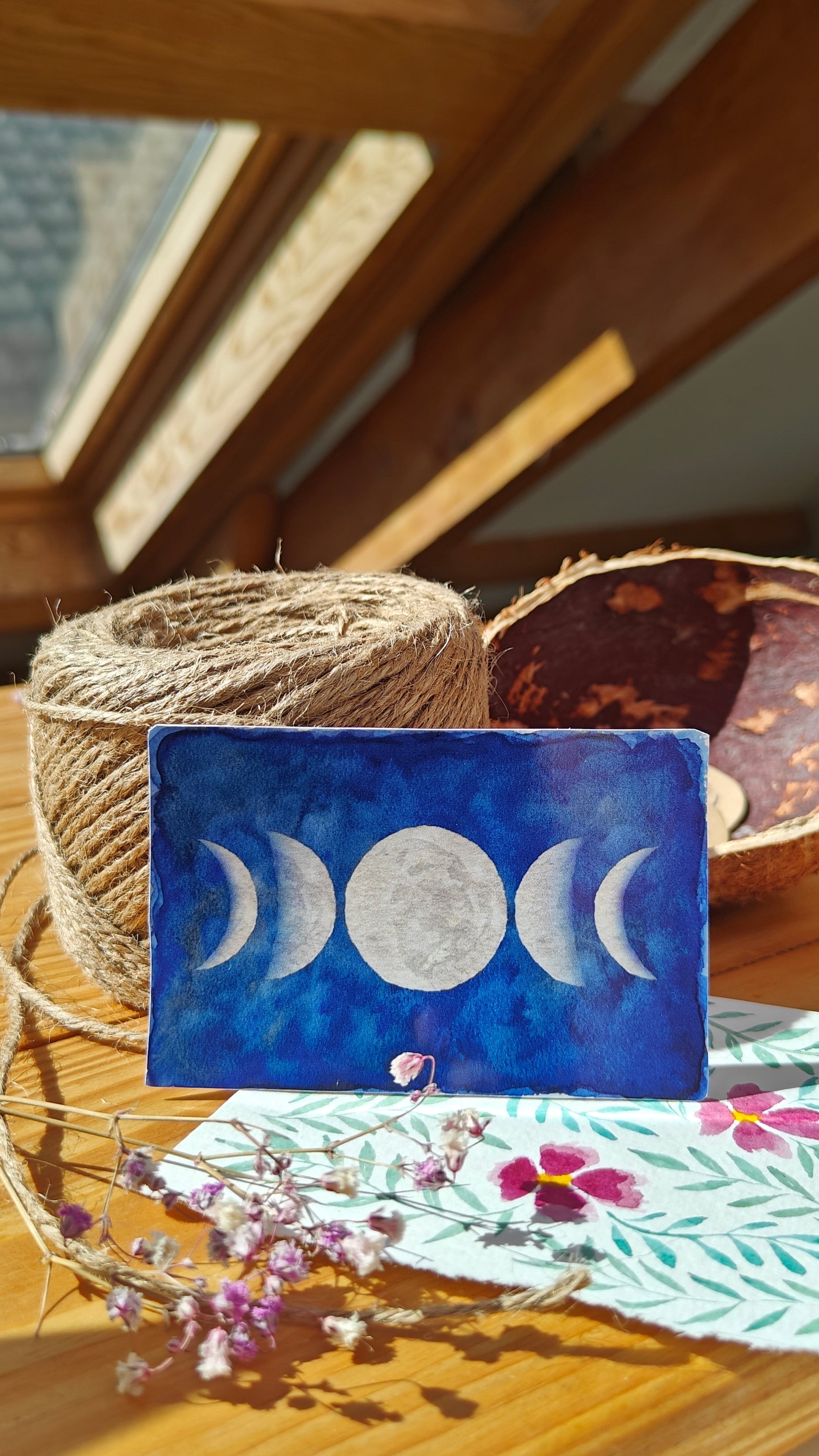 Sticker with moon phases over a wooden surface with twine, watercolour painting and flowers.