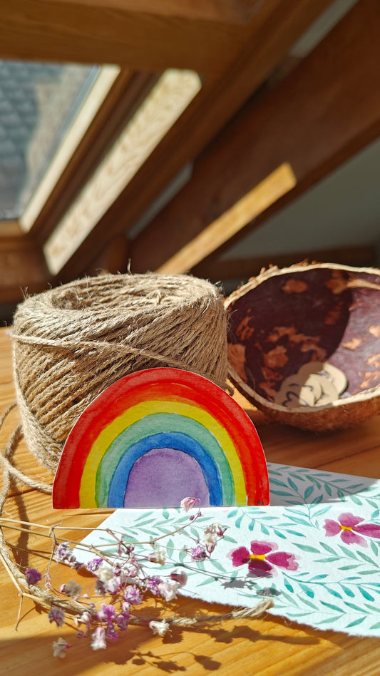 Colourful watercolour rainbow-shaped sticker on a wooden table, with twine, flowers, other natural elements and painting