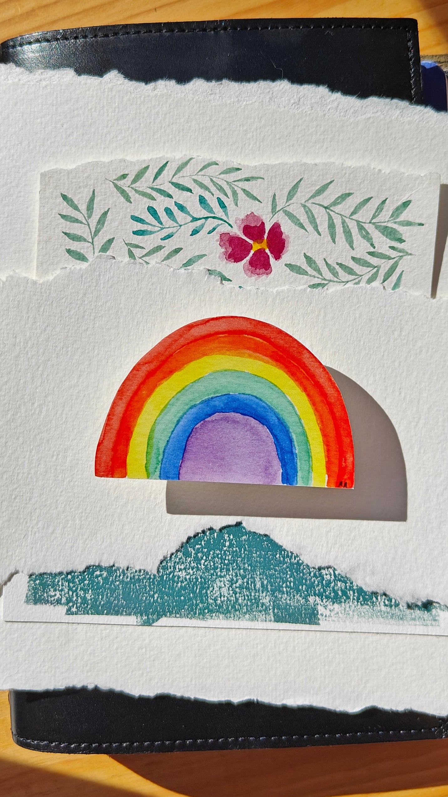 Colourful watercolour rainbow-shaped sticker on hand painted pieces of paper, and a leather-covered journal and wooden surface.