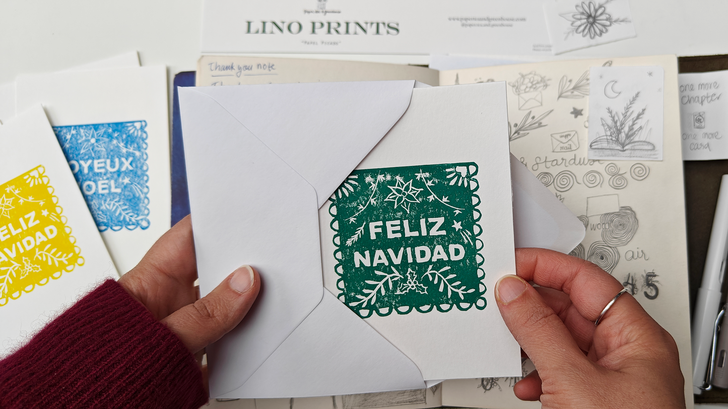Hand holding a card with 'Feliz Navidad' Mexican Papel Picado, hand card lino print, surrounded by stationery items on a table.
