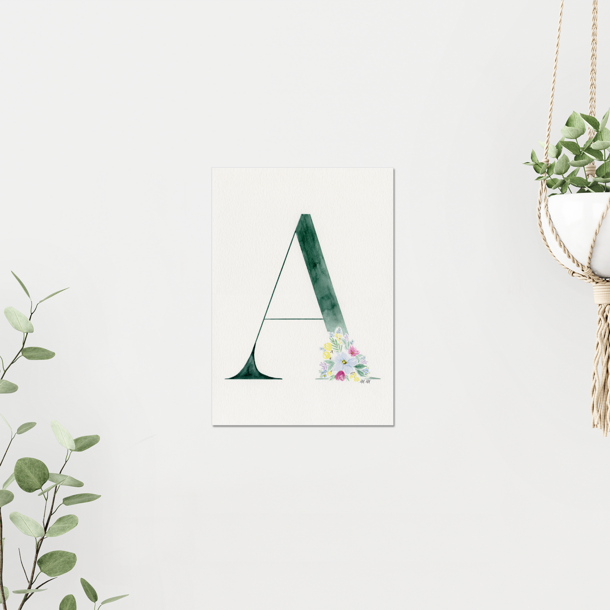 Decorative letter 'A' print with floral design on a white wall, surrounded by greenery. Made with watercolour.