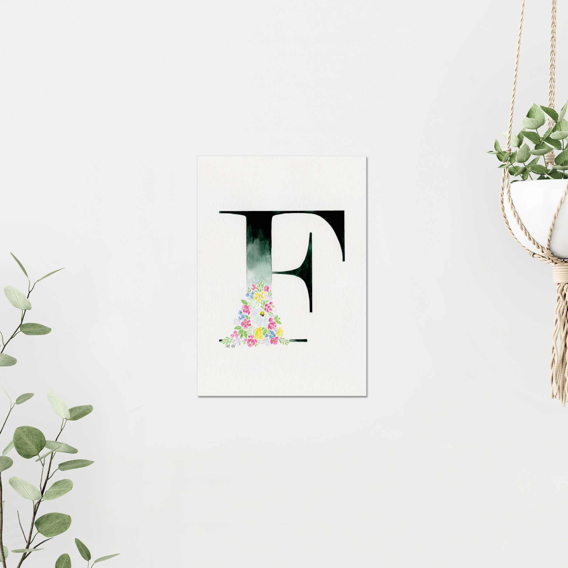 Decorative letter 'F' with floral design on a white background, on white wall surrounded by plants. Made with watercolours.