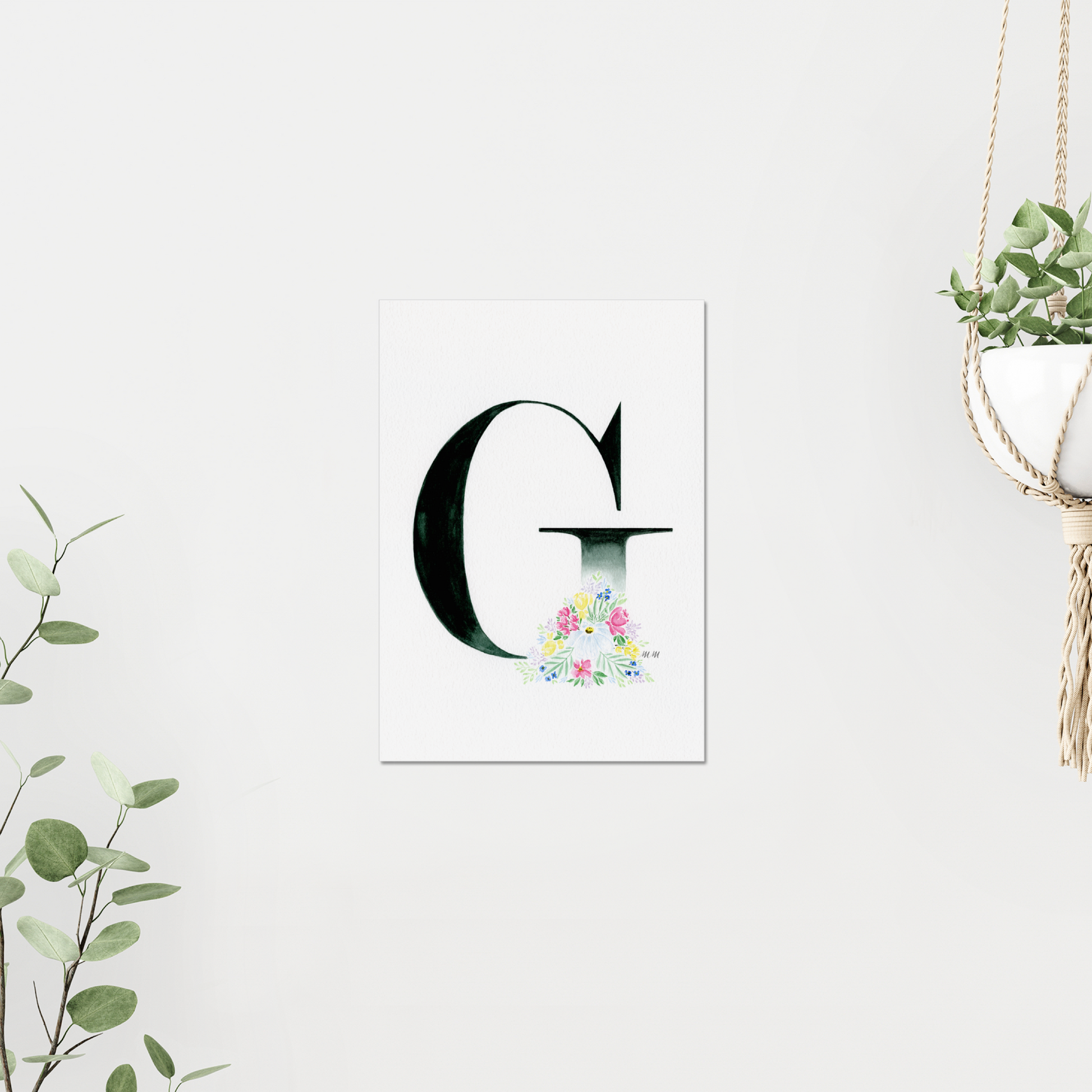 Decorative letter 'G' with floral design on a white background, on a white wall surrounded by plants. Made with watercolours.