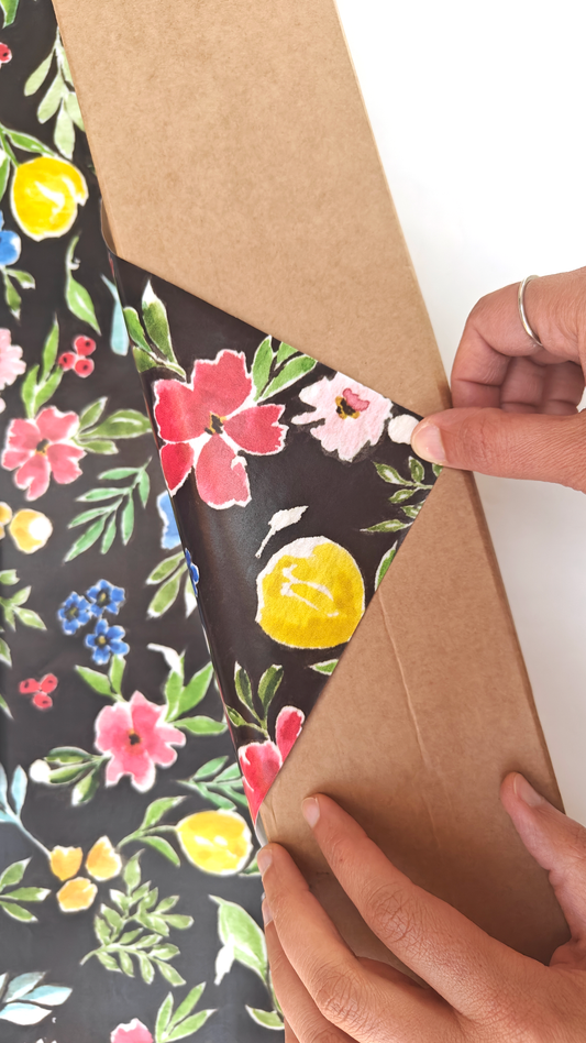 Person folding floral patterned wrapping paper, colourful flowers on a black background, over a brown package. Originally painted in watercolours.