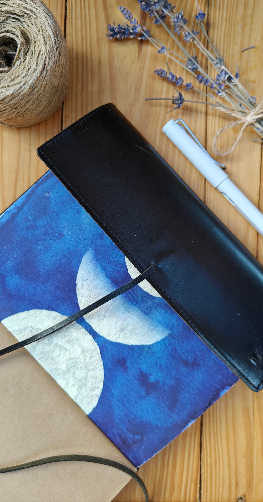 Blue sky and moon cycle watercolour designed journal in a leather cover on a wooden table with a pen, twine and lavender.