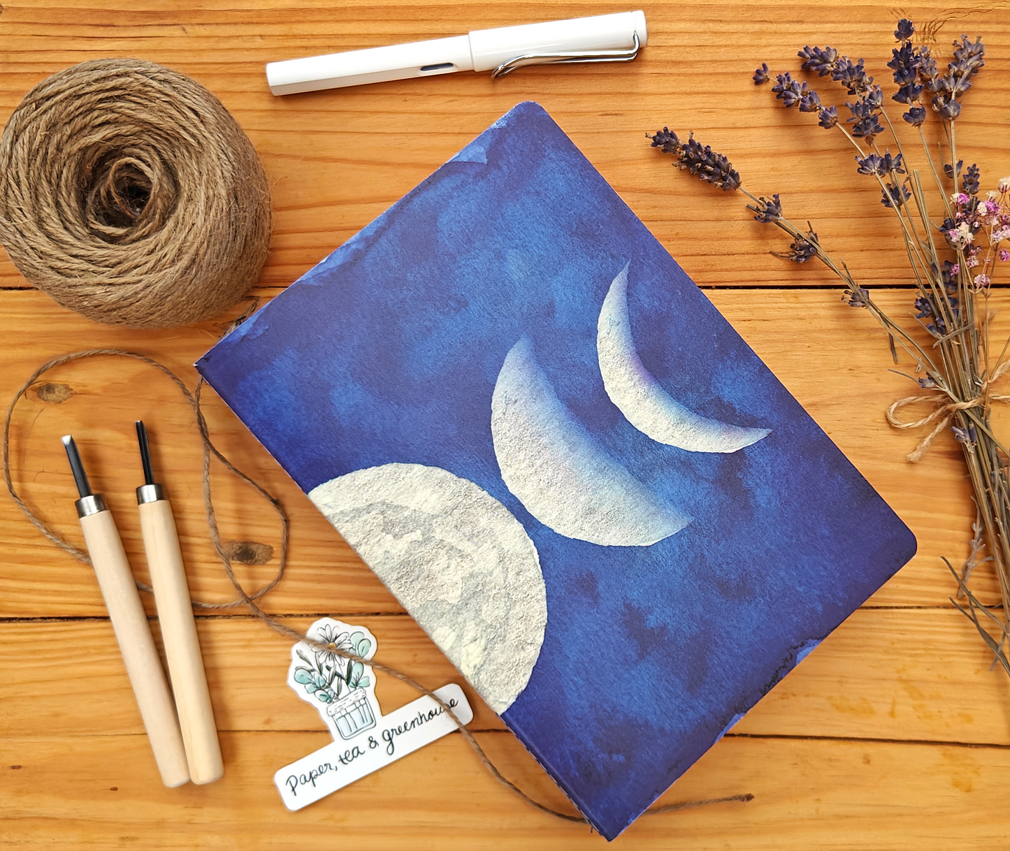 Blue notebook with moon design with watercolour look, on a wooden surface with twine, carving tools, a white pen, and lavender.