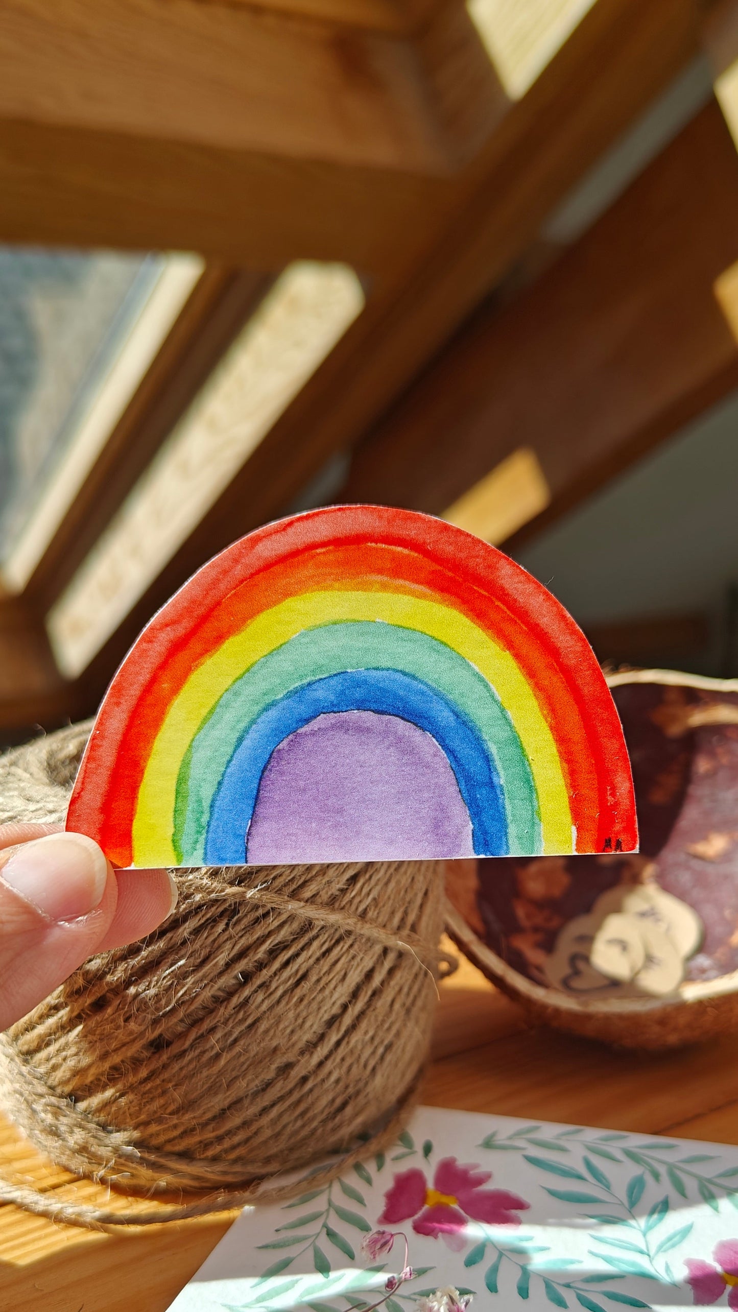 Hand holding a colourful watercolour rainbow-shaped sticker with a blurred background with twine, flowers and painting