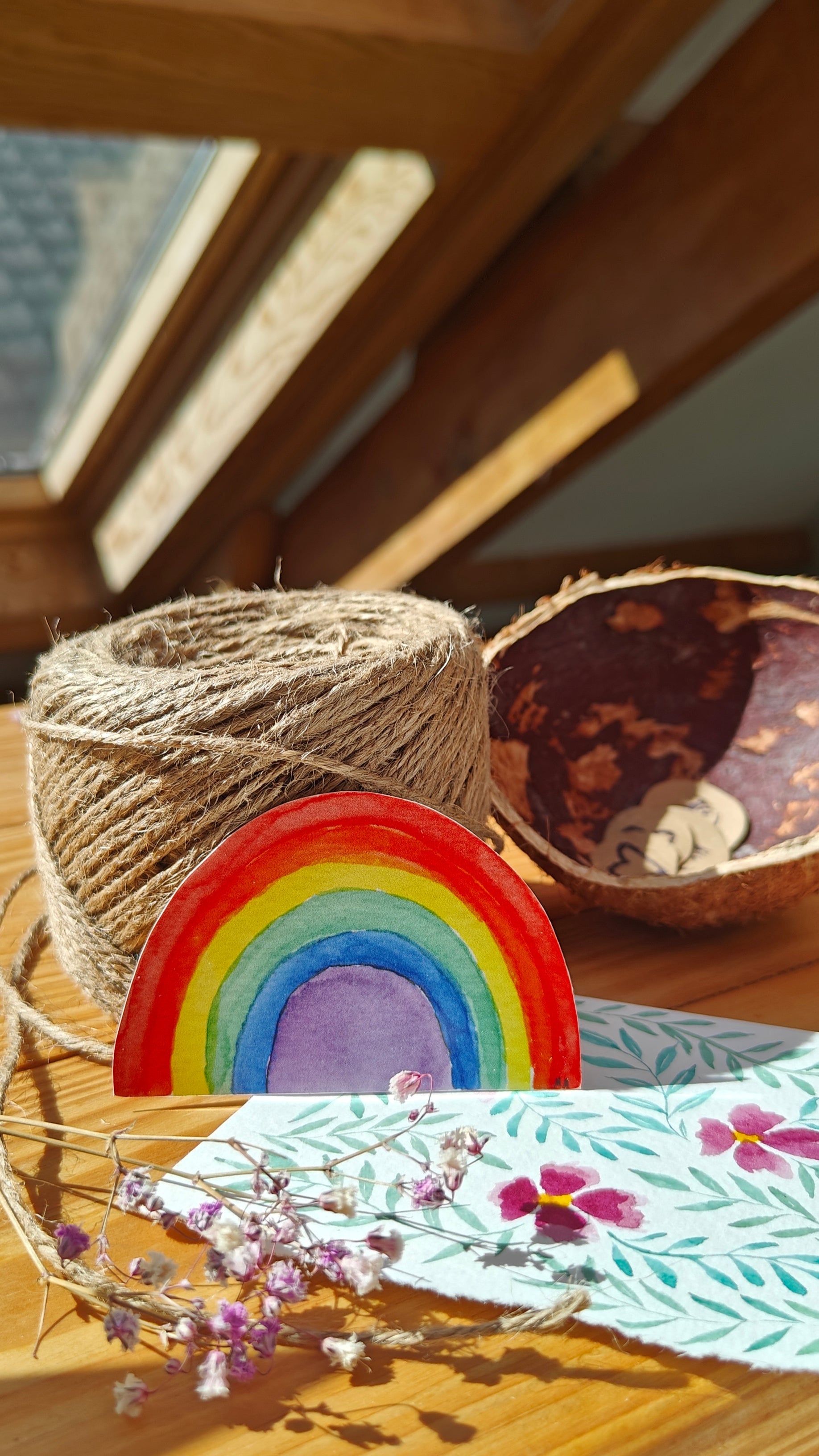 Colourful watercolour rainbow-shaped sticker on a wooden table, with twine, flowers, other natural elements and painting