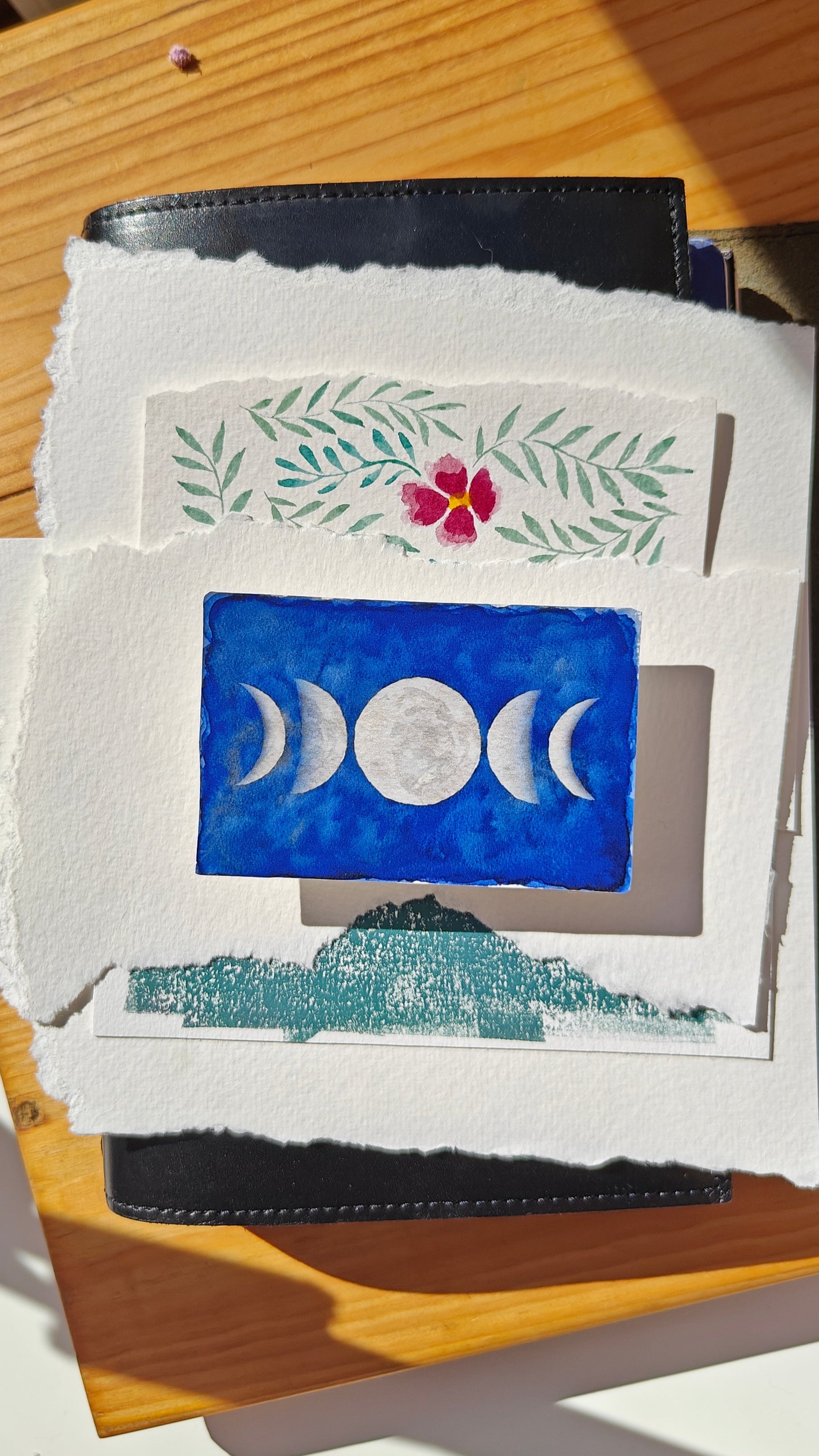 Sticker of a watercolour painting of the different moon phases, over various cuttings of paper and a leather journal