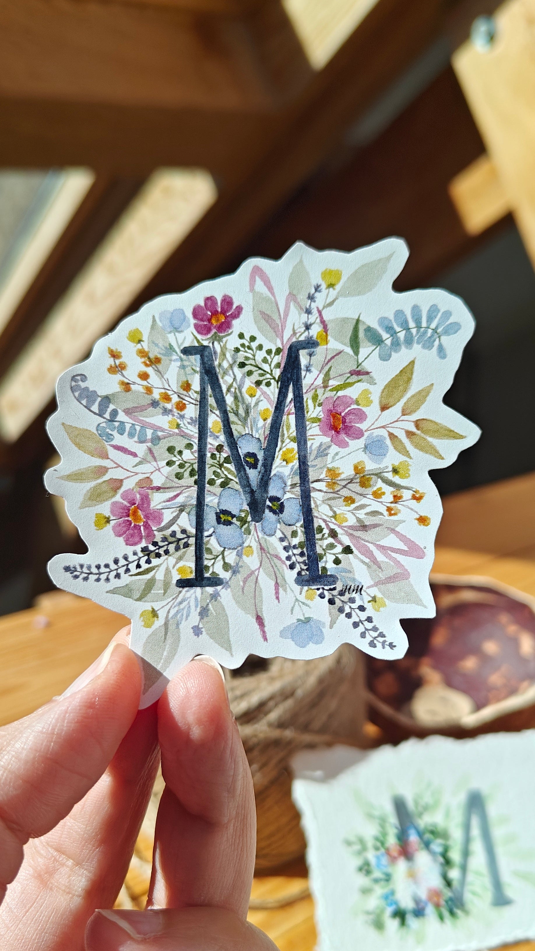 Decorative sticker with floral design and letter 'M' held by a hand on a wooden surface.