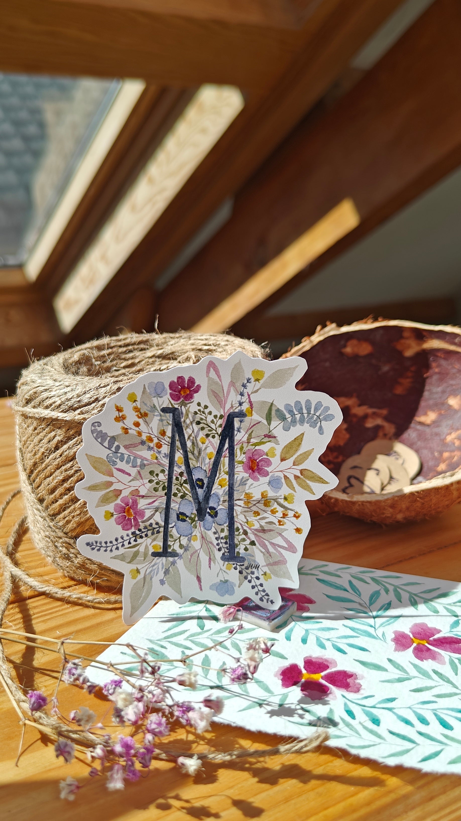 Decorative sticker with floral design and letter 'M' on a wooden surface with twine and flowers.