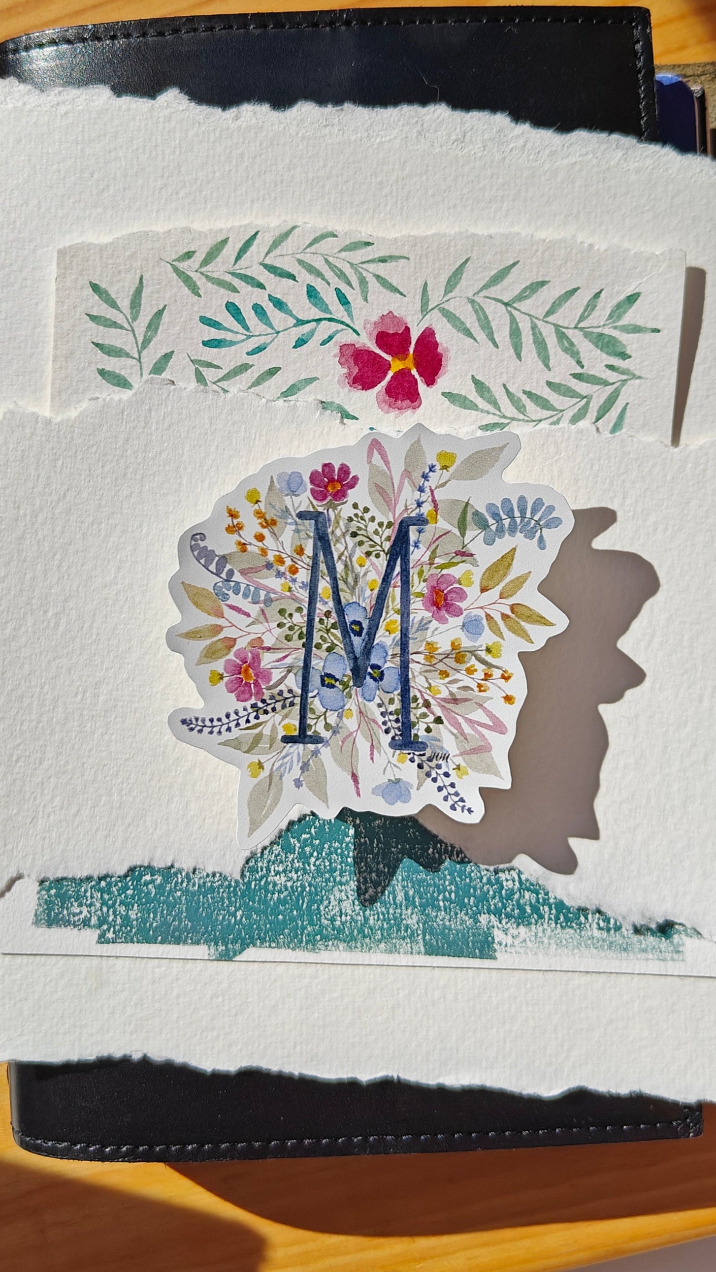 Decorative sticker of floral design with an 'M' on various decorated papers and a leather journal over a wooden surface