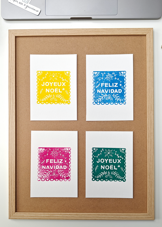 Four Mexican Papel Picado Lino prints with 'Feliz Navidad' and 'Joyeux Noel' text in different colours on a wooden frame.