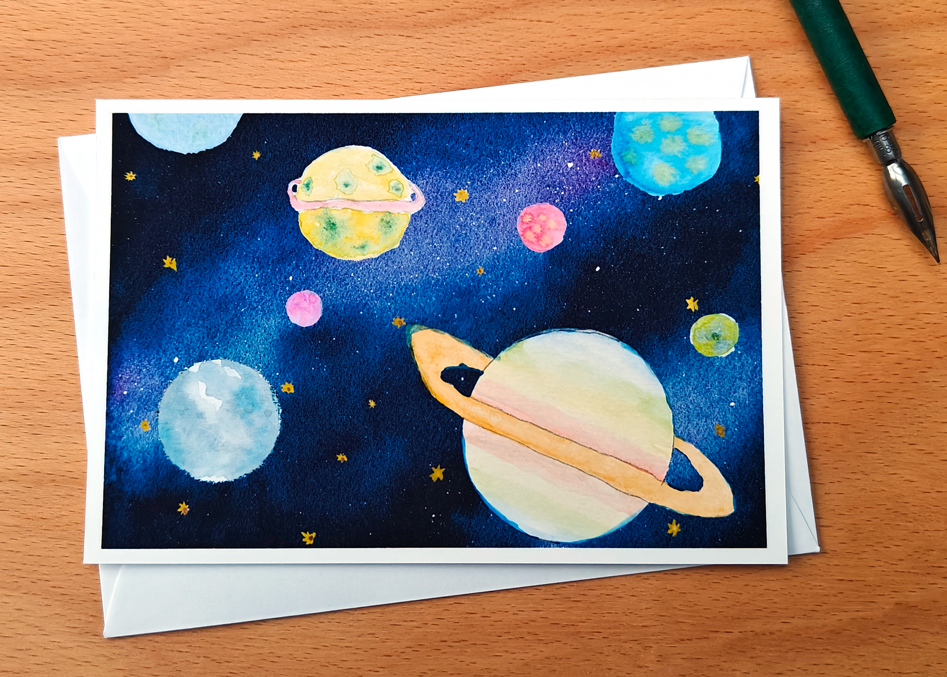 Card with space-themed illustration of planets painted with watercolours, with an envelope and a pen on a wooden surface