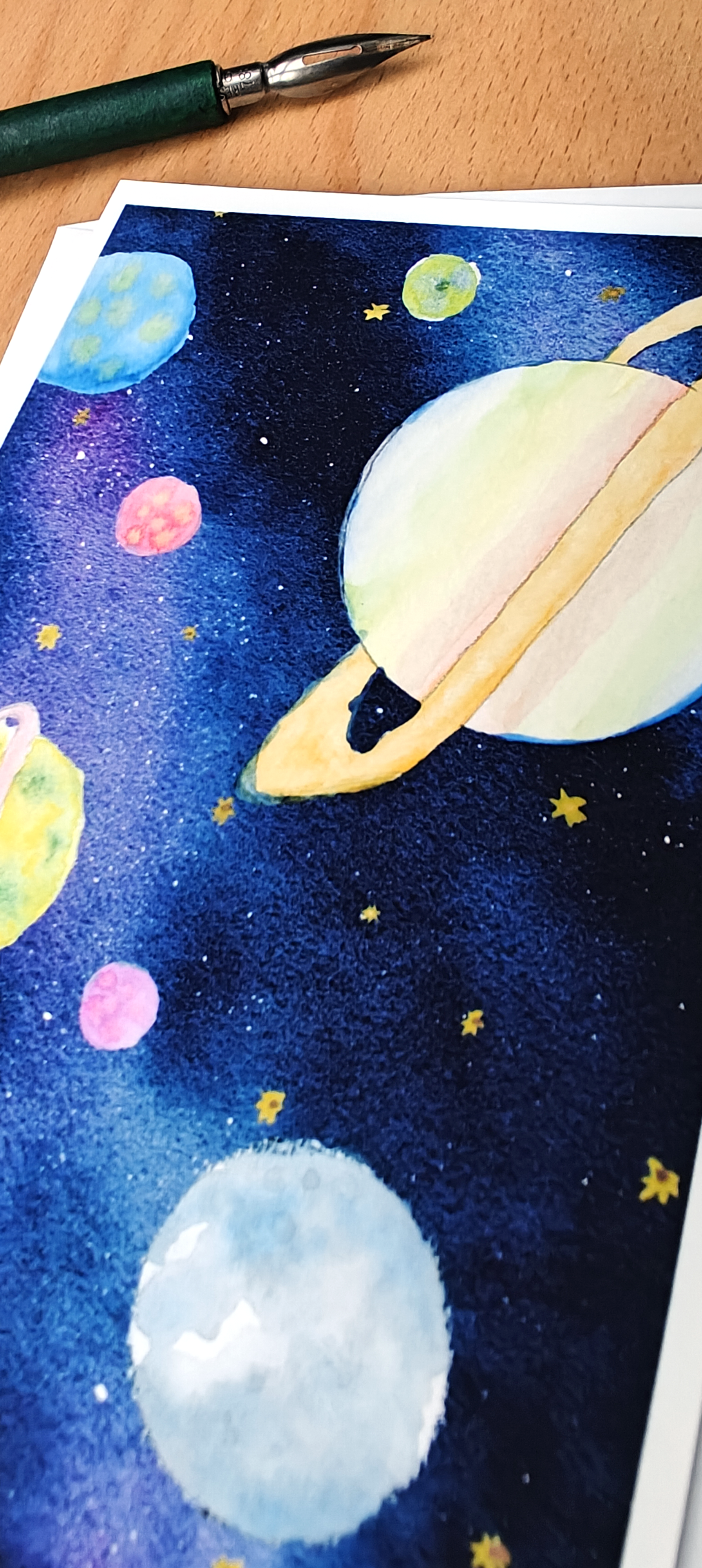 Space-themed artwork with planets and stars on a piece of paper, with a pen at the top.