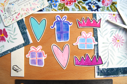 Colourful stickers of hearts, gift boxes, and crowns on a wooden surface with floral paintings.