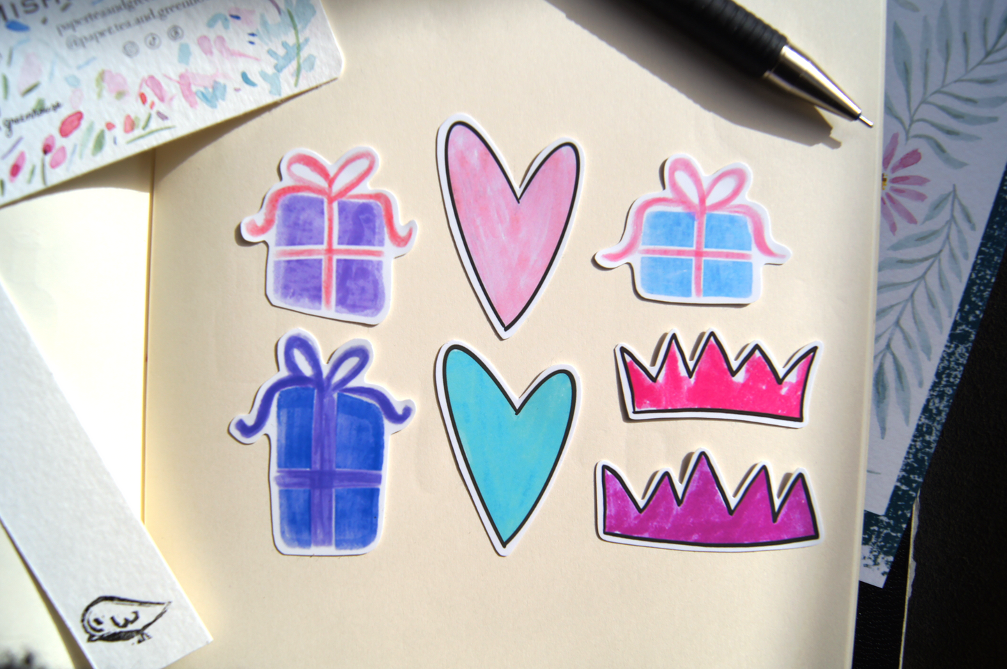 Colourful stickers of presents, crowns and hearts on a journal, with a watercolour painting, a bookmark, and a pencil.