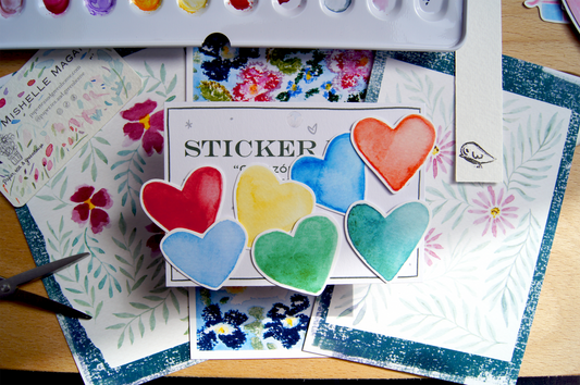 Pack of 7 heart-shaped stickers, all in different colours over stacked postcards, scissors and a watercolour palette.