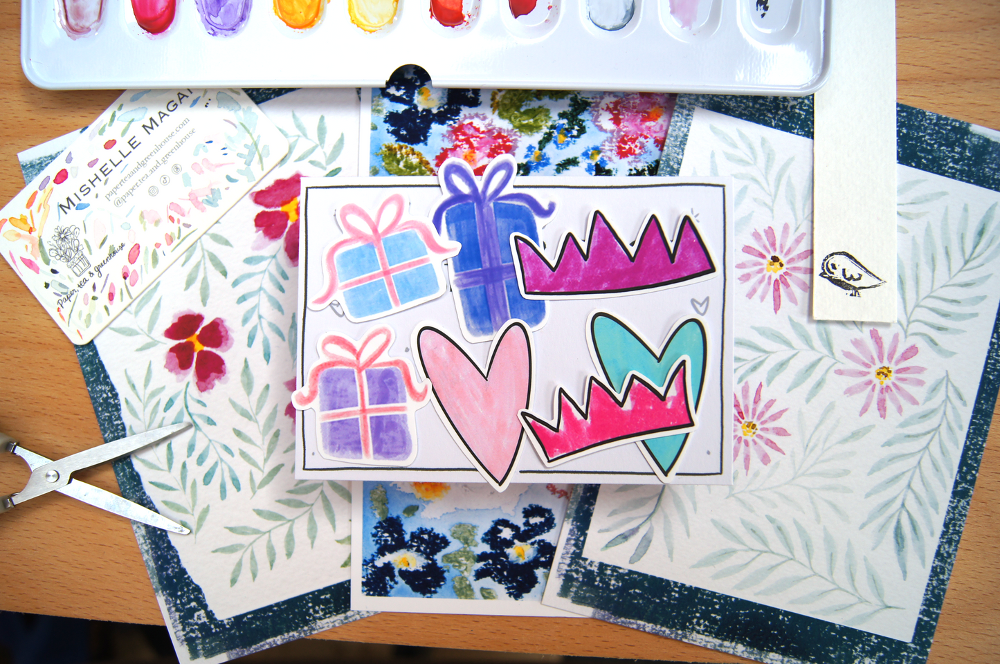 Colourful stickers with art supplies including watercolour paints, and paper with artistic designs on a wooden surface.