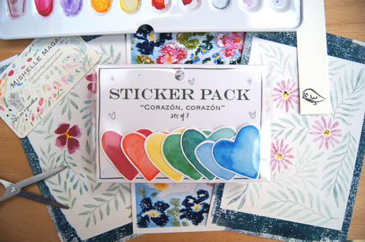 Pack of 7 heart-shaped stickers, all in different colours over stacked postcards, scissors, a handmade bookmark with a bird, and a watercolour palette.