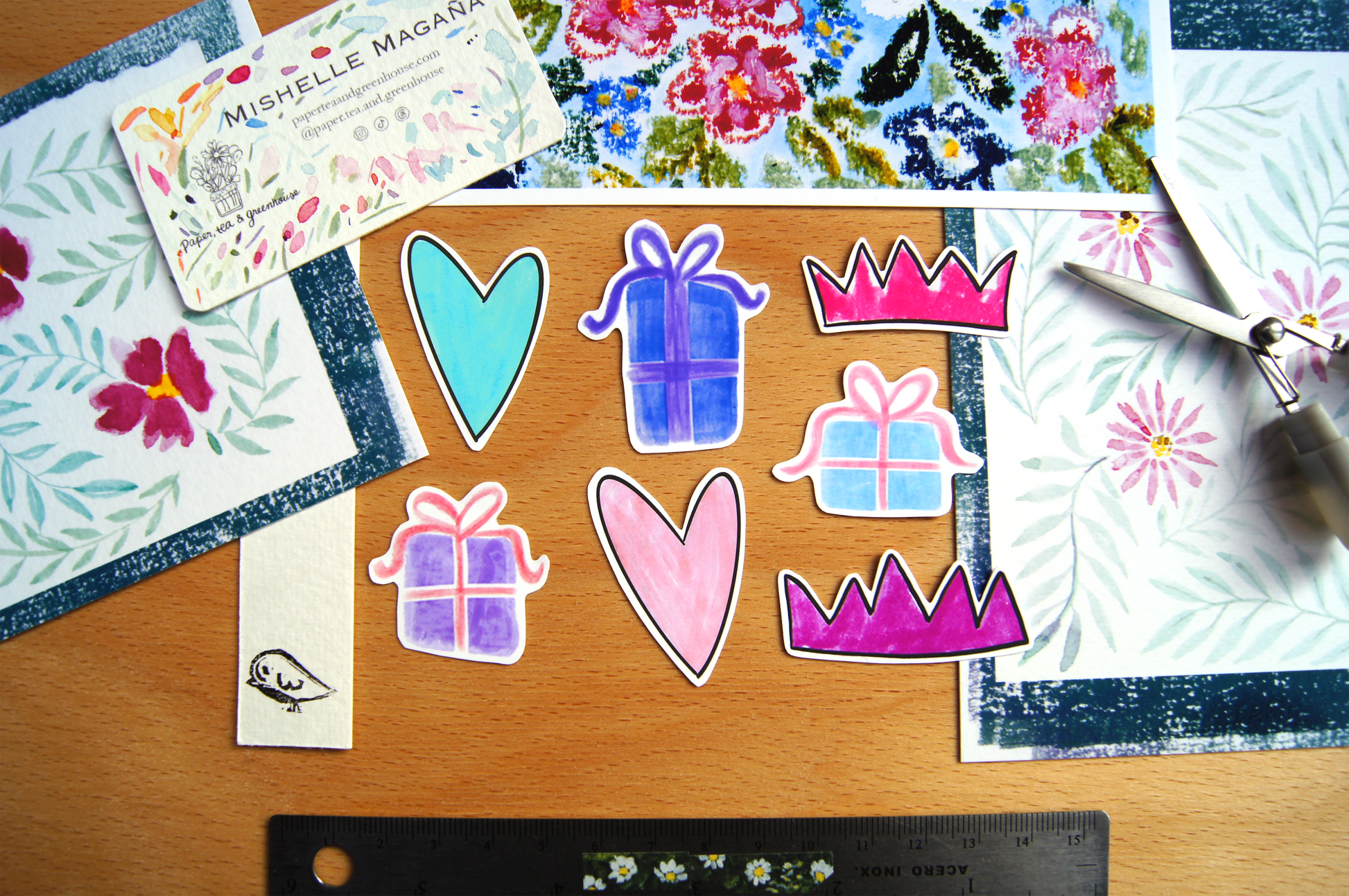 Colourful stickers of hearts, gifts, and crowns on a wooden surface with floral watercolour paintings, a bookmark with a lino-print bird and scissors in the background.