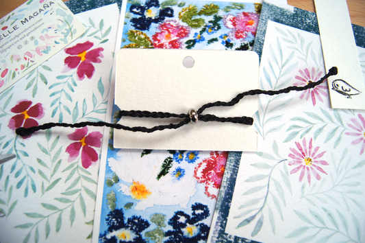 Back of bracelet with adjustable bead, in its original package with a background floral watercolour paintings.