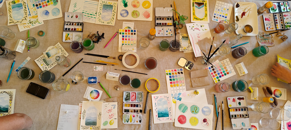 Art supplies including watercolor paints, brushes, and paper on a table.