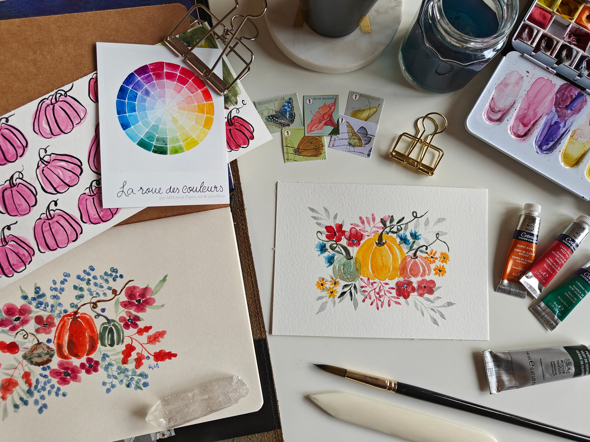 A styled flat-lay of watercolor painting supplies and finished artwork on a white surface, including a color wheel card, painted pumpkin illustrations, a floral watercolor painting, paint tubes, brushes, clips, and a watercolor palette.