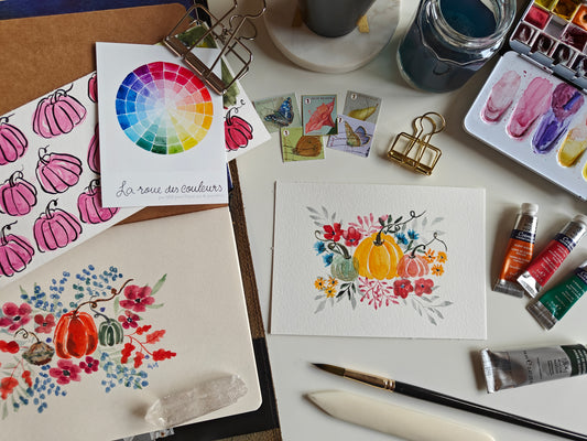 A styled flat-lay of watercolor painting supplies and finished artwork on a white surface, including a color wheel card, painted pumpkin illustrations, a floral watercolor painting, paint tubes, brushes, clips, and a watercolor palette.