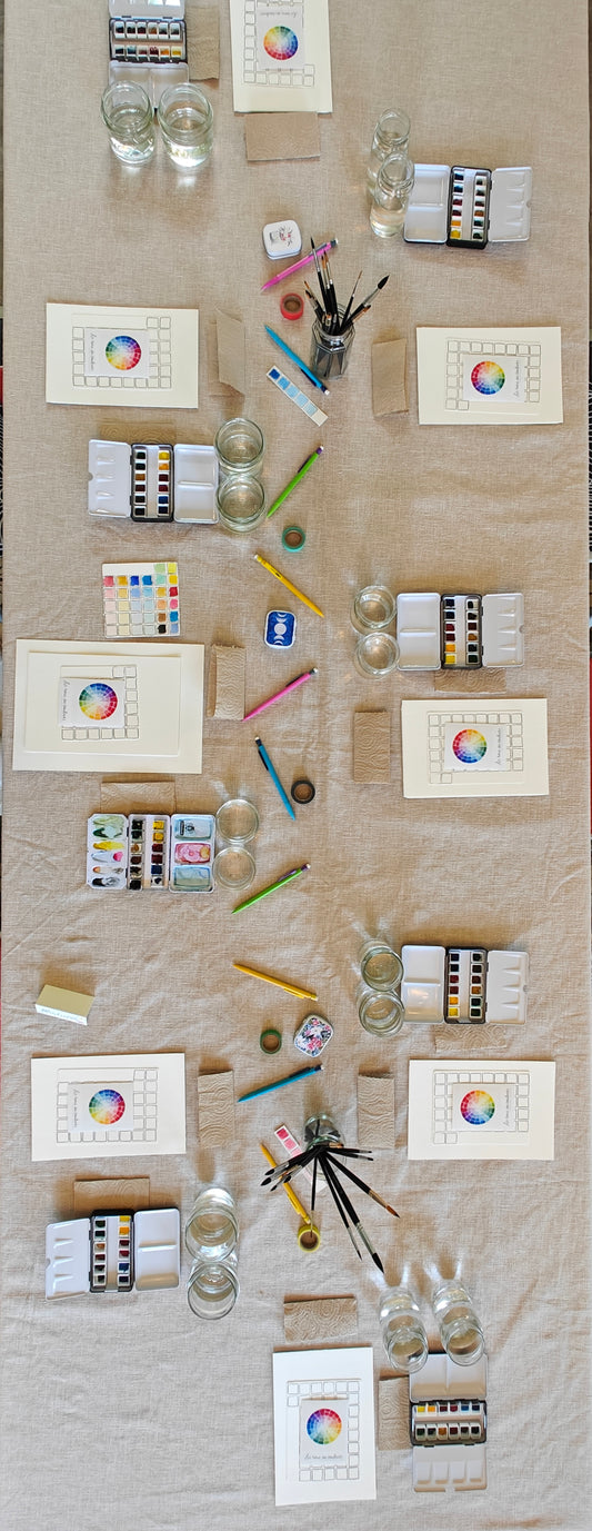 An art workshop setting including a collection of art supplies like paint tubes, brushes, a colour wheel, washi tape, pencils, and color swatches on a textured surface.