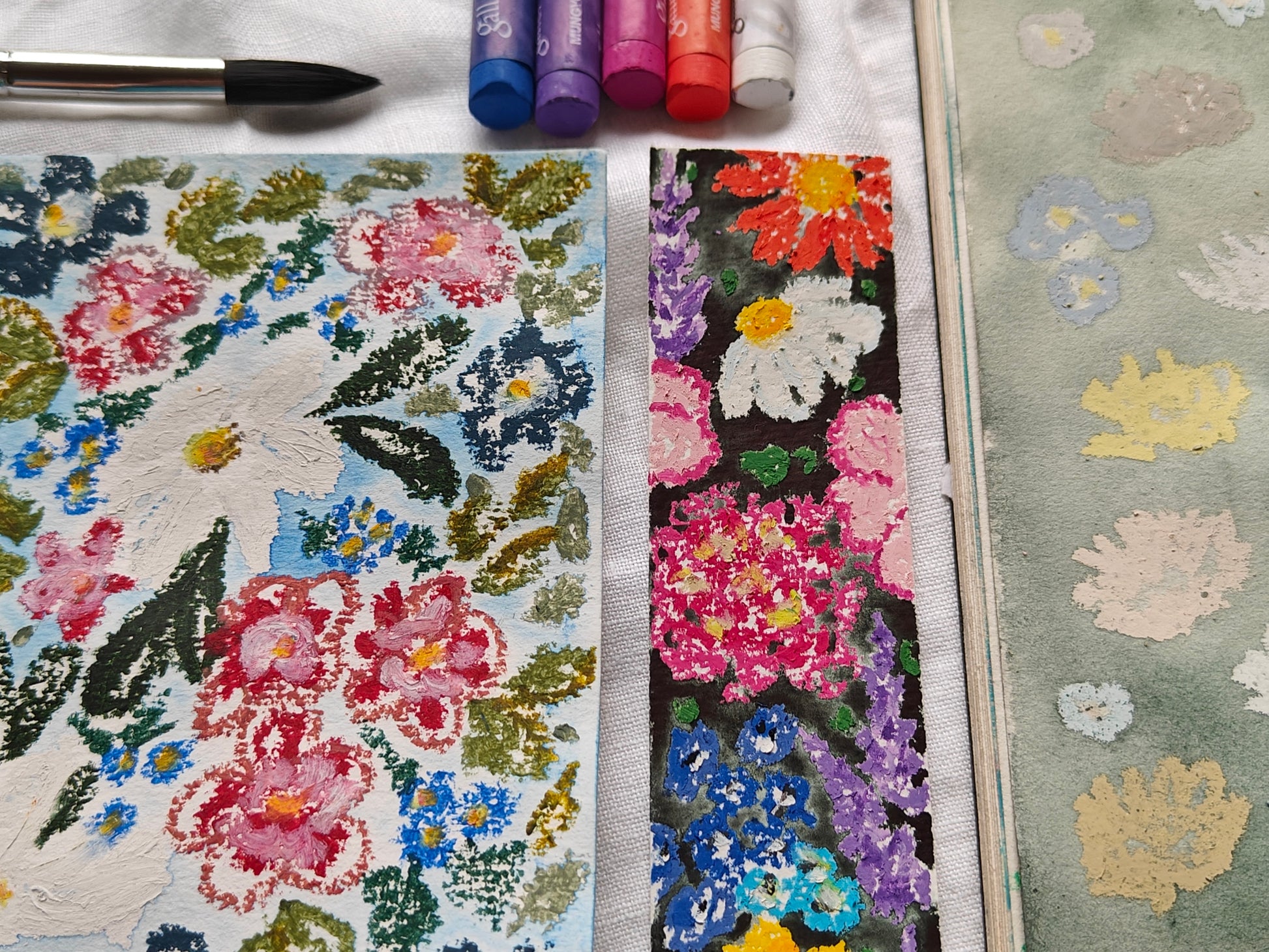 Two textured floral art cards or prints displayed side-by-side on a white fabric surface, with a paintbrush, oil pastels, and a green patterned card visible around them.