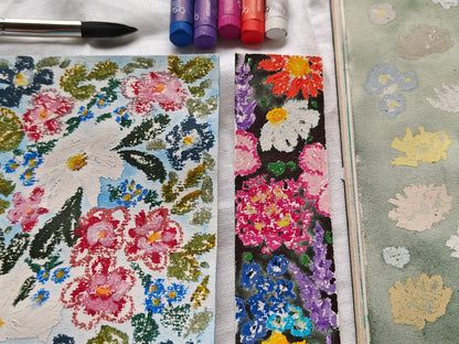 Two textured floral art cards or prints displayed side-by-side on a white fabric surface, with a paintbrush, oil pastels, and a green patterned card visible around them.
