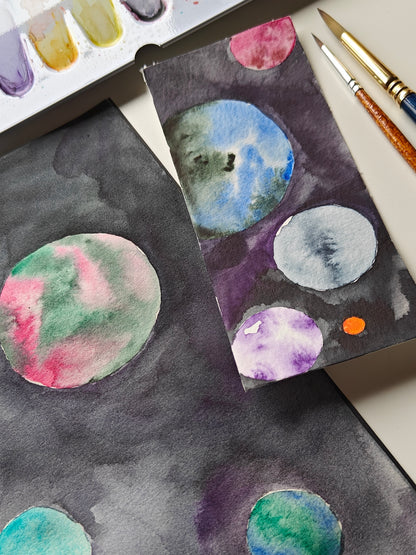 Watercolor painting on paper showing abstract circular planets or moons in various colors like blue, green, pink, purple, and gray on a dark space-like background, with paintbrushes and a paint palette partially visible.