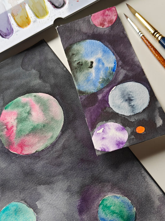 Watercolor painting on paper showing abstract circular planets or moons in various colors like blue, green, pink, purple, and gray on a dark space-like background, with paintbrushes and a paint palette partially visible.