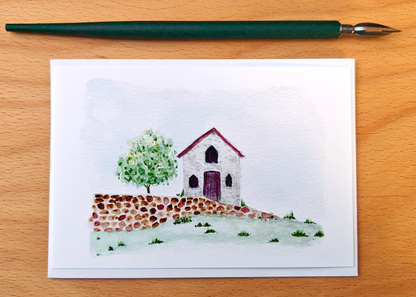 Small watercolour painting of a cottage and tree on a card, placed on a wooden surface.