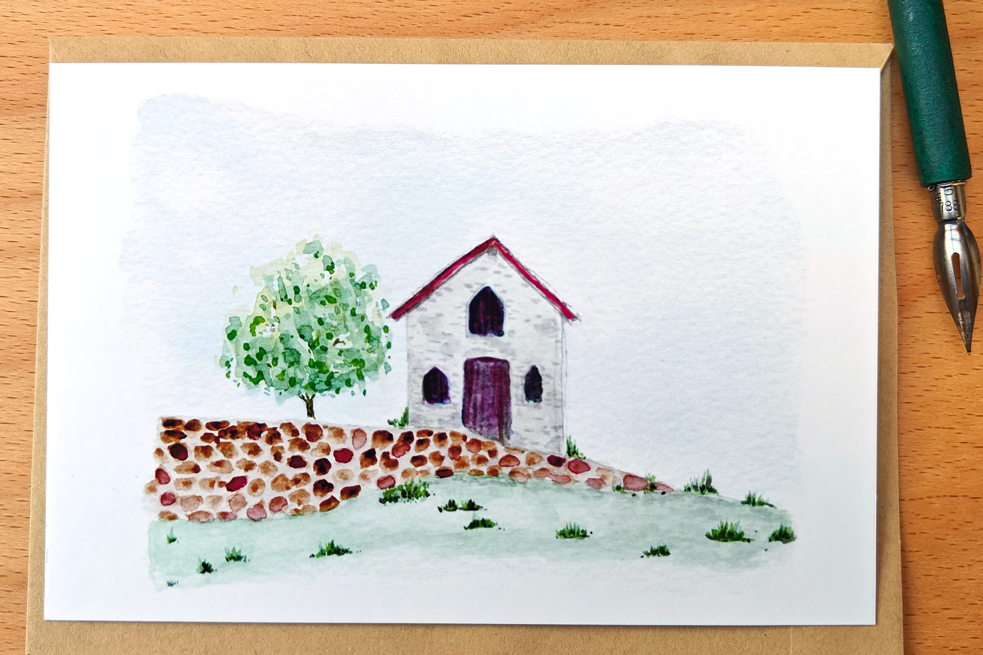 Hand-painted house and tree on a card with a pen on a wooden surface