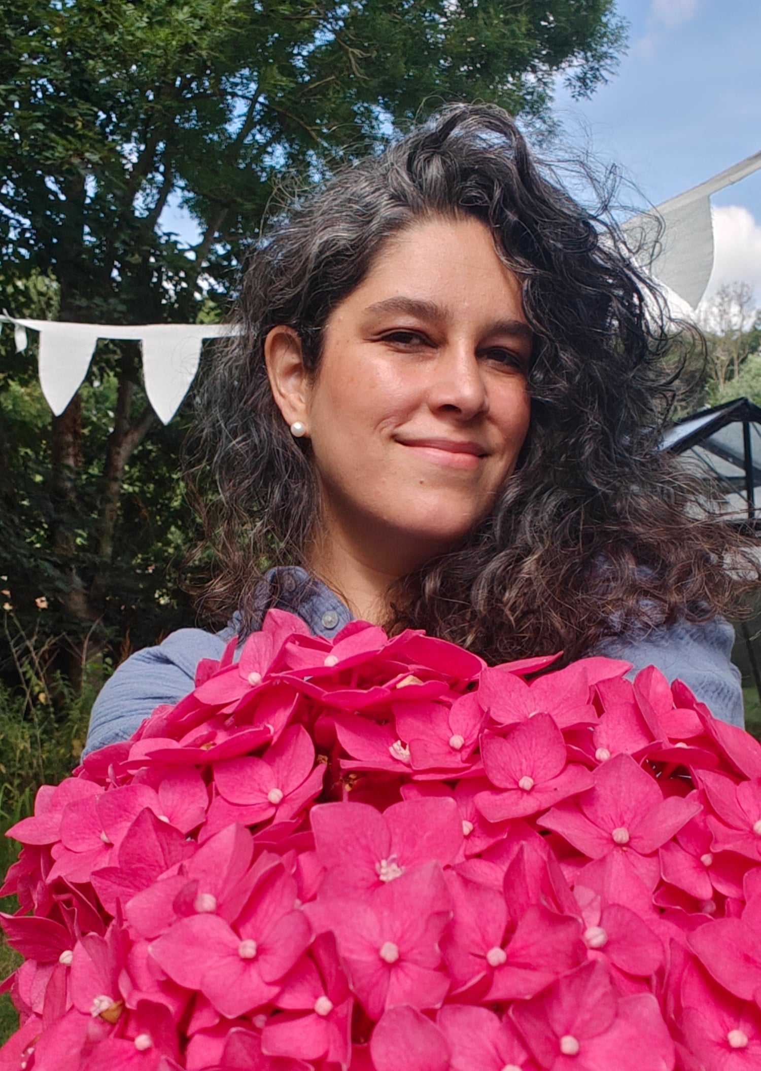 Mishelle Magaña, woman holding a large bouquet of pink flowers outdoors with greenery and blue sky in the background.