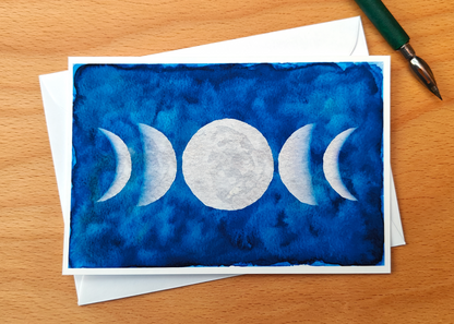 Post Card with moon phases design on a blue watercolour background, placed on a wooden surface with a pen.