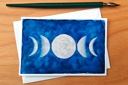 Greeting card with moon phases design on a blue watercolor background, placed on a wooden surface with a pen.