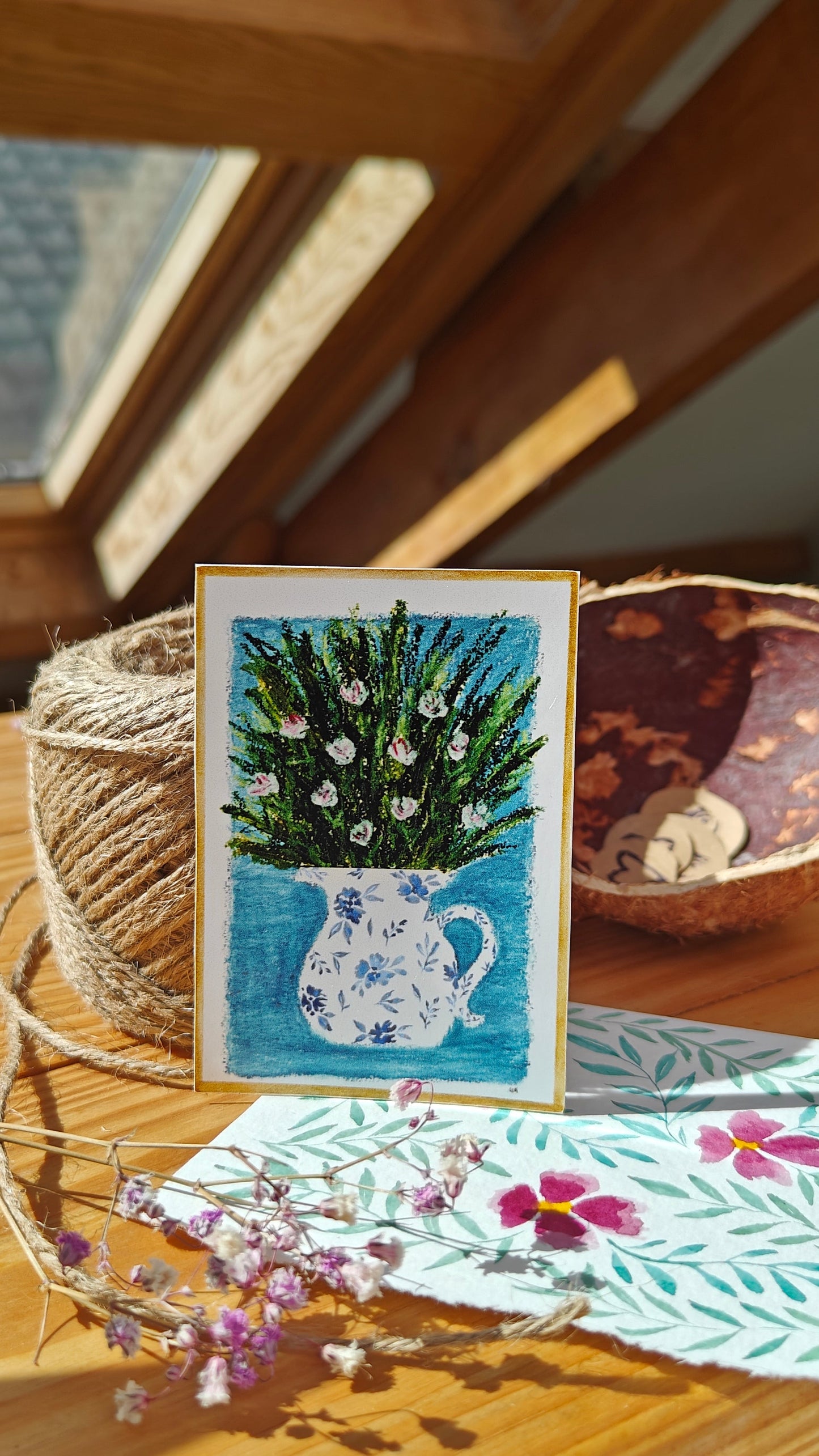 Sticker of an oil pastel and watercolour painting of a decorated, white pitcher with flowers and greenery on wood surface surrounded by natural objects