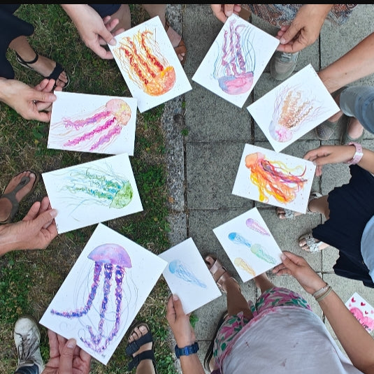 A group of people holding up colorful watercolor drawings of jellyfish in a circle outdoors on a paved path with grass.