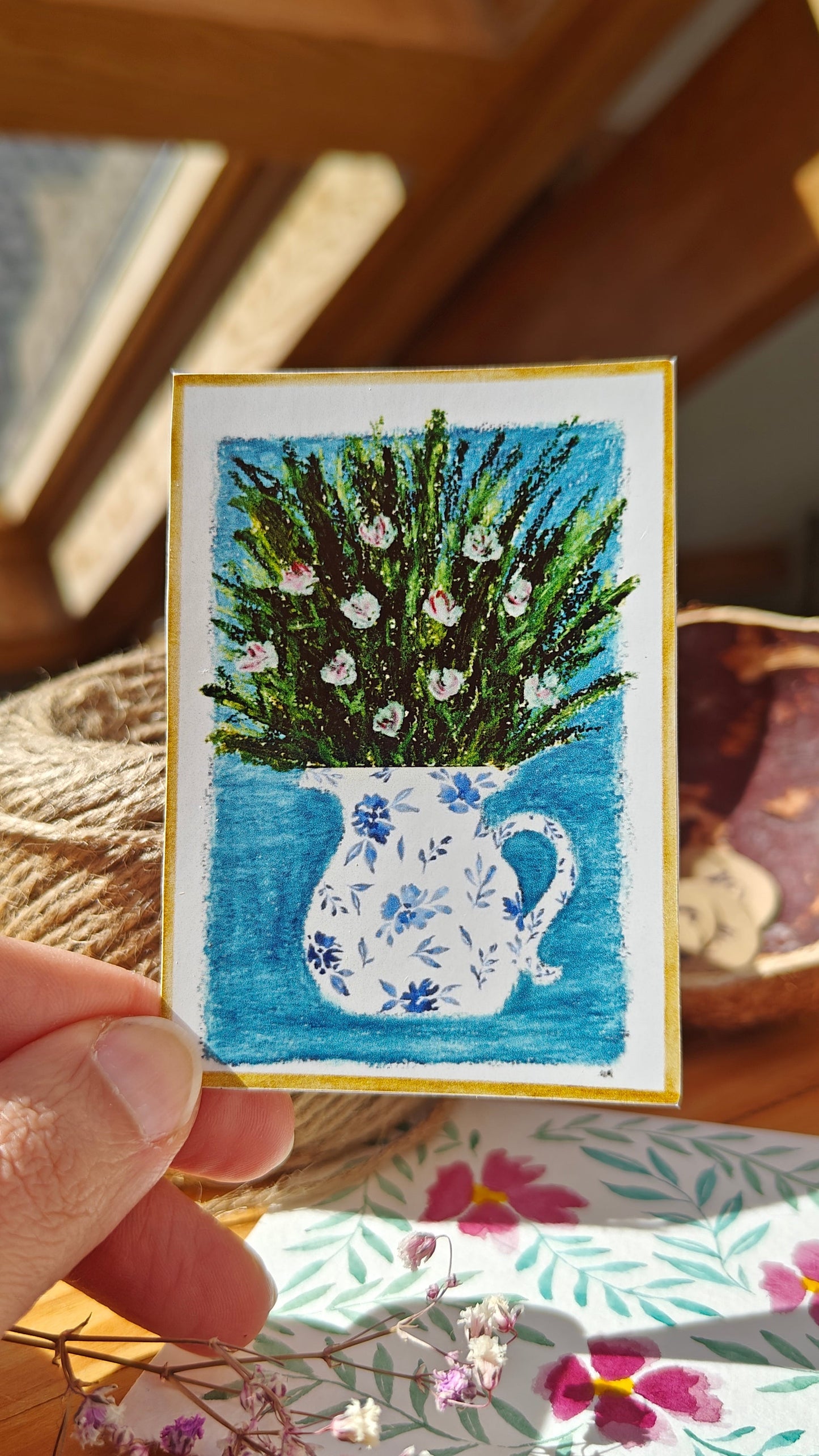 Sticker of an oil pastel and watercolour painting of a decorated, white pitcher with flowers and greenery held by a hand