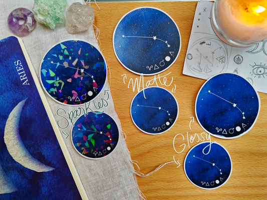 Set of blue astrological Constellation Stickers on a wooden surface with a candle, a journal, sketches, tarot cards and crystals.