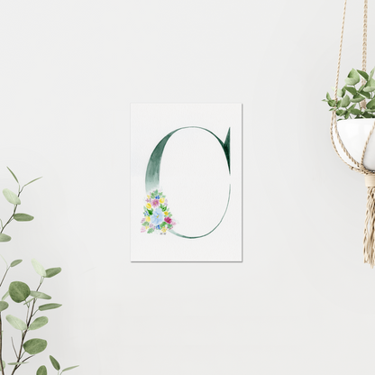 Decorative letter 'C' with floral design on a white background, surrounded by greenery. On white wall surrounded by plants. Made with watercolours.